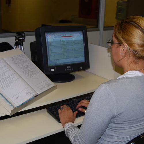 Une femme travaille à son ordinateur avec un livre ouvert à côté d'elle. Référence photographique : Mia Harbitz